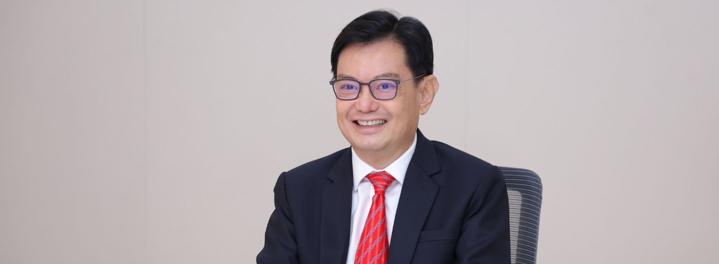 Man in suit, tie, and glasses smiling, seated against a light background.